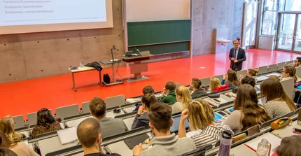 Ein Mann steht in einem Hörsaal vor vielen Studierenden und hält eine Vorlesung