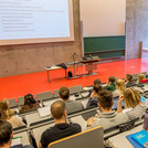 Ein Mann steht in einem Hörsaal vor vielen Studierenden und hält eine Vorlesung