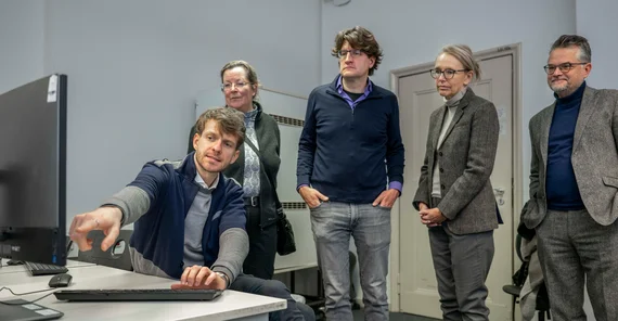 Mehrere Personen stehen vor einem PC-Arbeitsplatz. Eine Person gibt Erläuterungen was auf dem Bildschirm zu sehen ist.