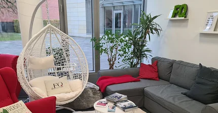 Kleine Sitzecke der FQ-Lounge: Rechts steht ein Sofa mit roten und schwarzen Kissen, davor ein runder weißer Couchtisch mit Flyern und Zeitschriften. Ein roter Sessel ist zum Tisch ausgerichtet, daneben hängt ein weißer Hängekorb mit Polstern. Dahinter steht ein zweiter roter, nach links gedrehter Sessel. Im Hintergrund eine Fensterwand mit Pflanzen.