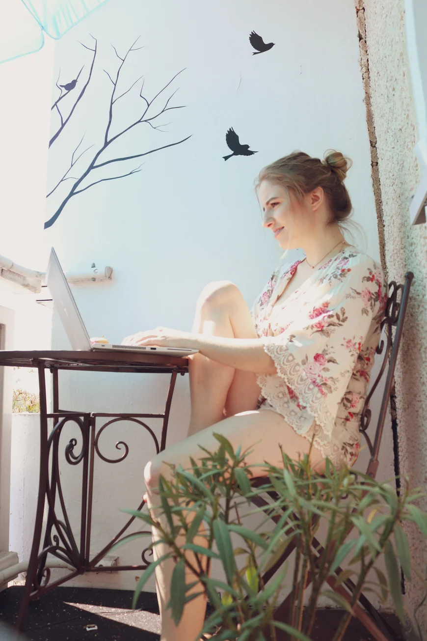 Eine Frau sitzt im Sommerkleid bei sonnigem Wetter auf dem Balkon an ihrem Laptop. Auf der Wand sind zwei Vogelsilhouetten angebracht, vor ihr steht eine Topfplanze
