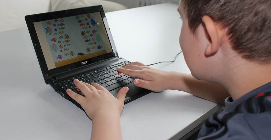 A child is sitting at a table and is looking to a tablet. The fingers are laying on the keyboard.