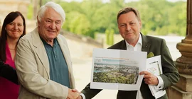 Two men shake hands, one holds a printout of a building design up to the camera.