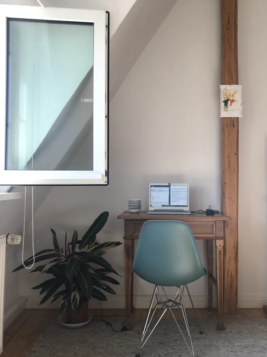 Ein SChreibtisch und eine Stehlampe neben einem geöffnenten Fenster. Auf dem Schreibtisch stht ein Laptop und neben dem Tisch eine Topfpflanze