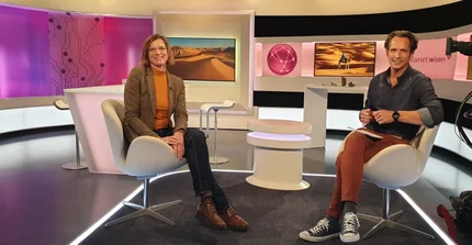 Prof. Linstädter and journalist sitting in a recording studio with cameras and screens with pictures of the desert in the background.