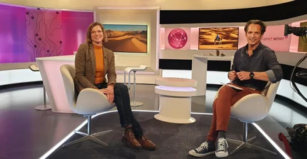 Prof. Linstädter and journalist sitting in a recording studio with cameras and screens with pictures of the desert in the background.