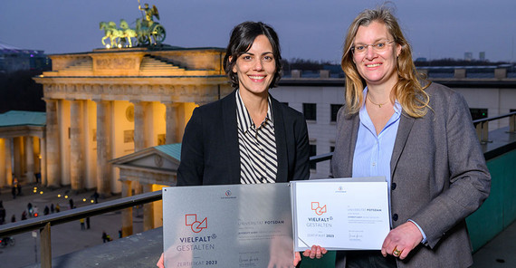 Dr. Britta van Kempen, Vizepräsidentin für Studium und Lehre, und Dr. Nina Khan, Referentin für Chancengleichheit und Diversity (v.r.n.l.) halten das Zertifikat „Vielfalt gestalten“. Im Hintergrund das Brandenburger Tor in Berlin.