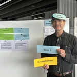 Eine männlich gelesene Person hält vor einer Moderationswand drei Moderationskarten hoch.