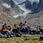 Menschen sitzen vor einer Berglandschaft, während einer vor ihnen steht und etwas erzählt