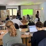 Das Foto zeigt eine Gruppe von Personen (Mitglieder des UFS) von hinten, die in verschiedenen Tischreihen sitzen und zwei Präsentierenden vorne zuhören. Die zwei präsentierenden Frauen zeigen eine PowerPoint-Präsentation.