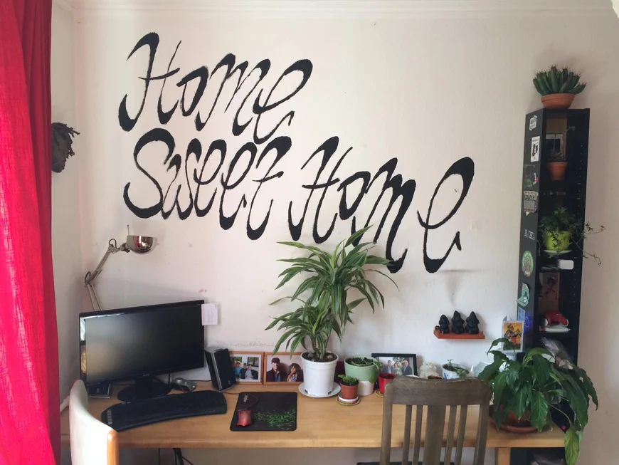 Auf der Wand hinter einem Schreibtisch steht in geschwungener Schrift "Home sweet home". Links auf dem Schreibtisch ein Bildschirm mit Tastatur, an der Wand entlang sind Bilderahmen mit Fotos und Topfpflanzen dekorativ angeordnet