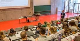 Ein Mann steht in einem Hörsaal vor vielen Studierenden und hält eine Vorlesung