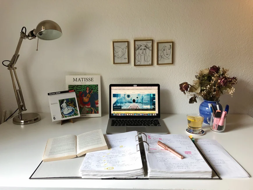 Schreibtisch mit Laptio, davor ein dicker Ordner und Schreibutensilien. An der Seit ein Kustkalender und ein Bildband von Matisse. An der Wand hängen 3 Skizzen: Eine Taube, Ein Frauengesicht, ein Frauengesicht mit Taube, vielleicht Picasso. Rechts daneben ein Blumenstraus und ein Glas Tee