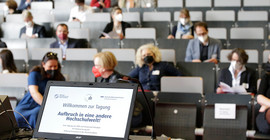 Das Bild zeigt einen Laptop an einem Rednerpult mit dem Tagungstitel "Willkommen zur Tagung. Aufbruch in eine andere Hochschulwelt!" und im Hintergrund das Publikum. Foto: Sandra Scholz