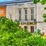 Durch Büsche gerahmter Blick auf das Portal eines großen Hauses, vor dem eine Rotkäppchenfigur steht