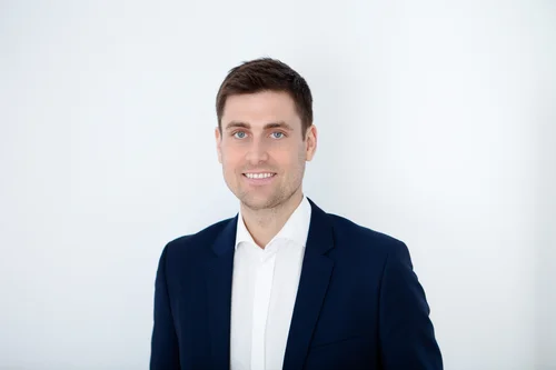 Ein Mann mit kurzen, dunklen Haaren, einem dunkelblauen Anzug und weißem Hemd darunter ist abgebildet. Der Hintergrund ist weiß.