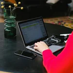 Eine Frau sitzt vor ihrem Laptop und arbeitet. Sie hat bestimmte Hilfsmittel neben dem Laptop liegen, um optimal arbeiten zu können mit ihrer Behinderung. Welche Behinderung bei der Frau vorliegt ist nicht sichtbar. 