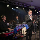 Ein Mann am Mikrofon, im Hintergrund Musiker am Schlagzeug und mit Gitarre.