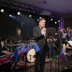 Ein Mann am Mikrofon, im Hintergrund Musiker am Schlagzeug und mit Gitarre.