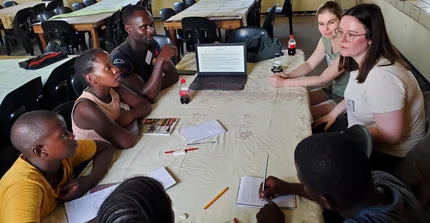 A mixed group of scientists and learners sitting around a table with an open laptop showing slides on water infiltration, notebooks and pencils on the table