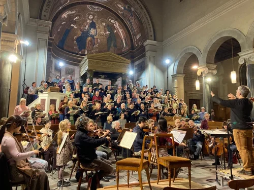 Ein Chor steht in einer Kirche, daneben einige Musiker mit Instrumenten, ganz rechts ein Dirigent