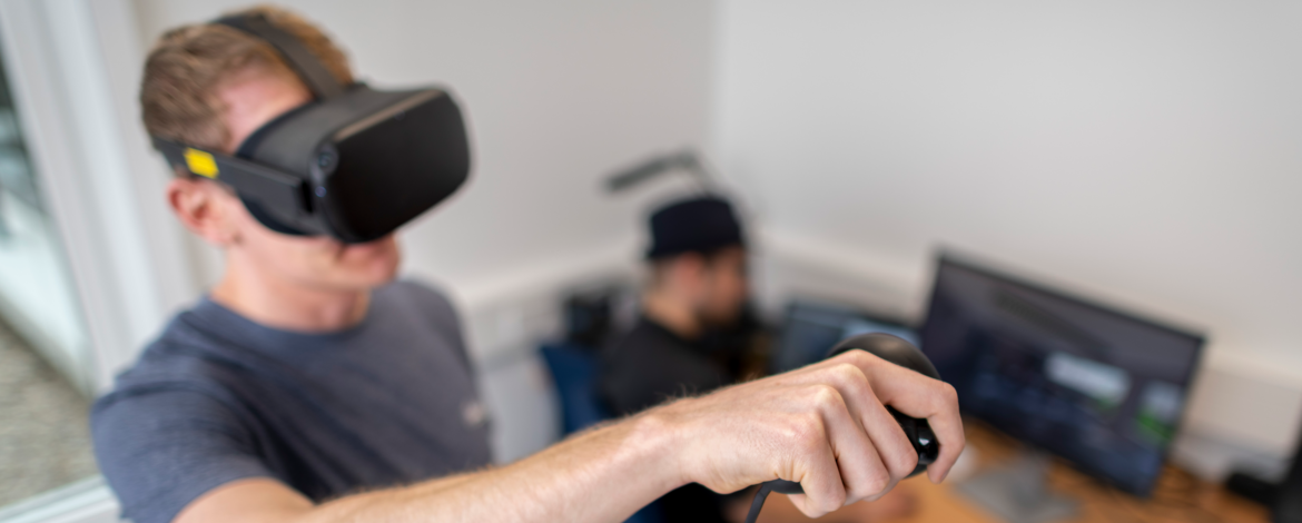 Eine Person mit VR-Brille auf dem Kopf, hat den Arm mit einem VR-Controller nach vorn ausgestreckt. Hinter der stehenden Person im Fokus, sitzt eie weitere Person vor einem Computer Bildschirm.