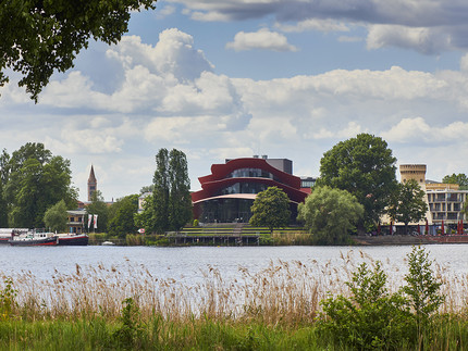 Man blickt über einen Fluss auf ein Theatergebäude mit rotem Dach