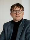 man with glasses and a gray jacket in front of a white wall