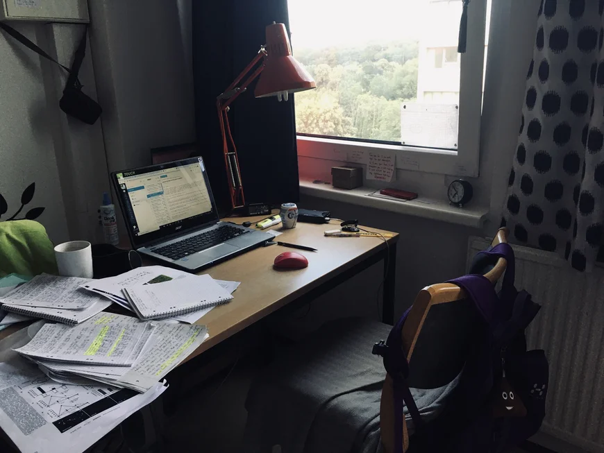 Schreibtisch mit Laptop und viel Papier. Seitlich Fenster mit Blick auf Wald