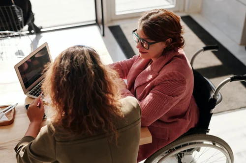 Zwei Personen sitzen an einem Tisch vor einem Laptop und unterhalten sich. Eine Person sitzt im Rollstuhl.