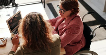 Zwei Personen sitzen an einem Tisch vor einem Laptop und unterhalten sich. Eine Person sitzt im Rollstuhl.