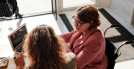 Zwei Personen sitzen an einem Tisch vor einem Laptop und unterhalten sich. Eine Person sitzt im Rollstuhl. 