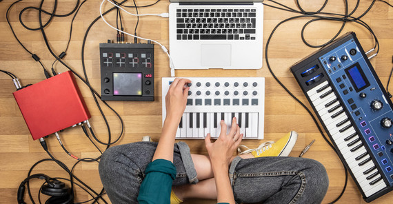 Person sitzt auf dem Fußboden im Schneidersitzt, umgeben von Keyboard, Laptop, Soundboard und anderen Geräten zum musizieren. Das Foto ist von AdobeStock/Photoboyko.