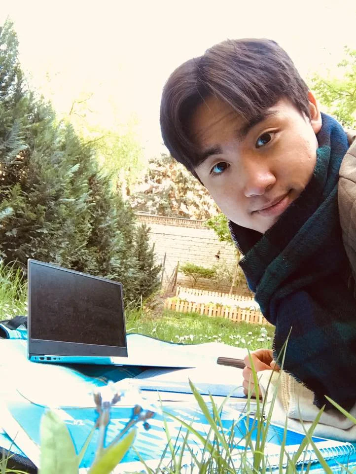 Ein junger Mann schaut in die Kamera, neben ihm auf einer Wiese steht der Laptop und Schreibutensilien liegen auf einer Decke