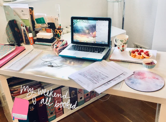 Kleiner Schreibtisch mit Stifbecher, einigen unterlagen und Notizbüchern, Laptio, etwas zu essen und Kaffeetasse. auf dem Bild steht "My weegend is all booked"