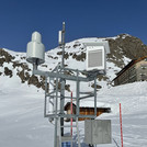 Field site Dresdner Hütte: Measuring equipment installed at a hillslope with snow. In the background is the chalet Dredner Hütte and a rocky mountaintop | Photo: Cosmic Sense Consortium
