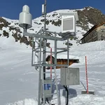 Field site Dresdner Hütte: Measuring equipment installed at a hillslope with snow. In the background is the chalet Dredner Hütte and a rocky mountaintop | Photo: Cosmic Sense Consortium