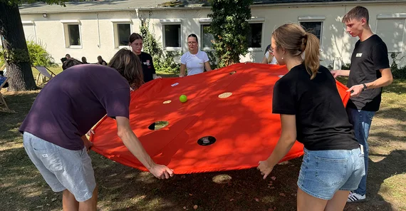 Teambuilding-Event mit dem Hochschulsport. Gemeinsam wird versucht, einen Ball nicht durch ein Loch in einer großen Plane rollen zu lassen, die mehrere Menschen zwwischen sich halten.
