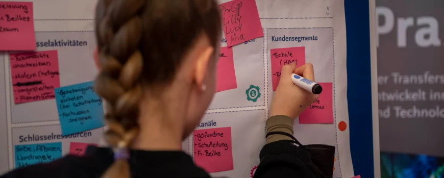 Schülerin steht vor einer Posterwand und pinnt ein Post it daran fest