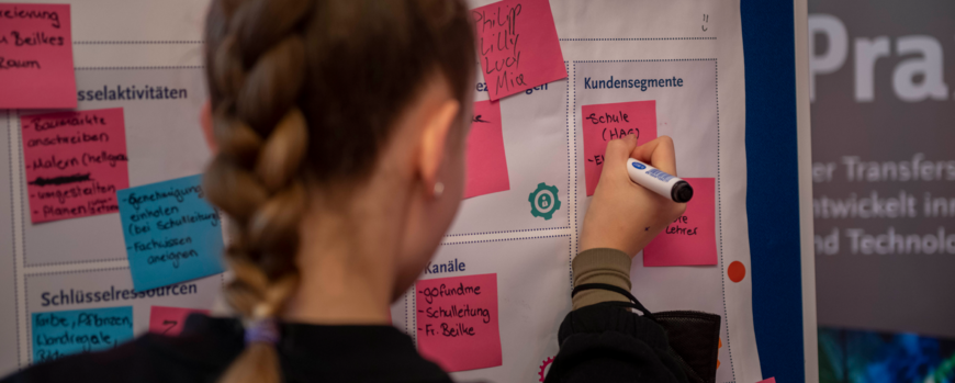 Schülerin steht vor einer Posterwand und pinnt ein Post it daran fest