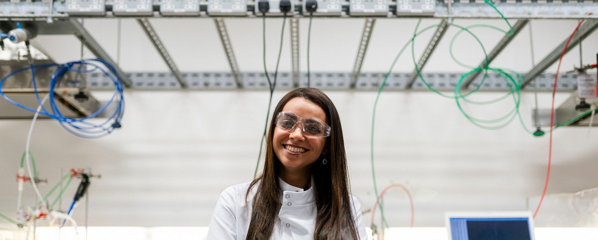 Eine weiße, junge, lächelnde Person mit langen braunen Haaren steht in einem Labor. Sie trägt einen weißen Laborkittel und trägt eine Laborbrile