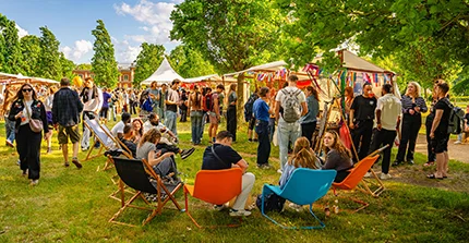 Eine Wiese mit Marktständen und vielen Menschen, im Vordergrund sitzen 4 Personen auf Liegestühlen mit dem Rücken zum Fotografen
