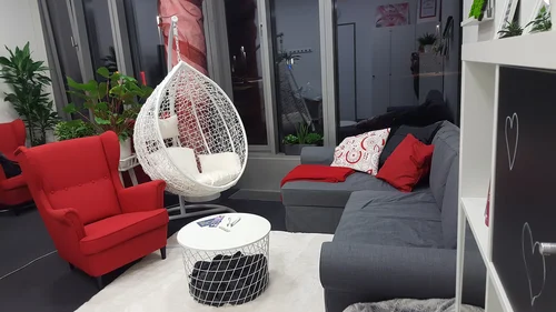 FQ-Lounge: Rechts steht ein weißes Regal mit schwarzen Türen und aufgemalten Herzen. Dahinter steht ein graues Sofa mit drei Kissen. Vor dem Sofa liegt ein weißer Teppich, auf dem ein runder, weißer Couchtisch in Korboptik steht, in dem eine graue Decke liegt. Links vom Tisch steht ein roter Sessel, daneben hängt ein weißer Hängekorb. Im Hintergrund eine Fensterwand mit Pflanzen.