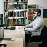 Ein Mann sitzt an einem Schreibtisch und arbeitet an einem Computer. Er trägt eine Brille mit dunklen Gläsern, da er sehbeeinträchtigt ist. 
