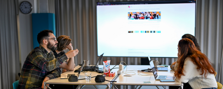 Vier Personen sitzen an einem Tisch, diskutieren miteinander und schauen auf eine Leinwand.