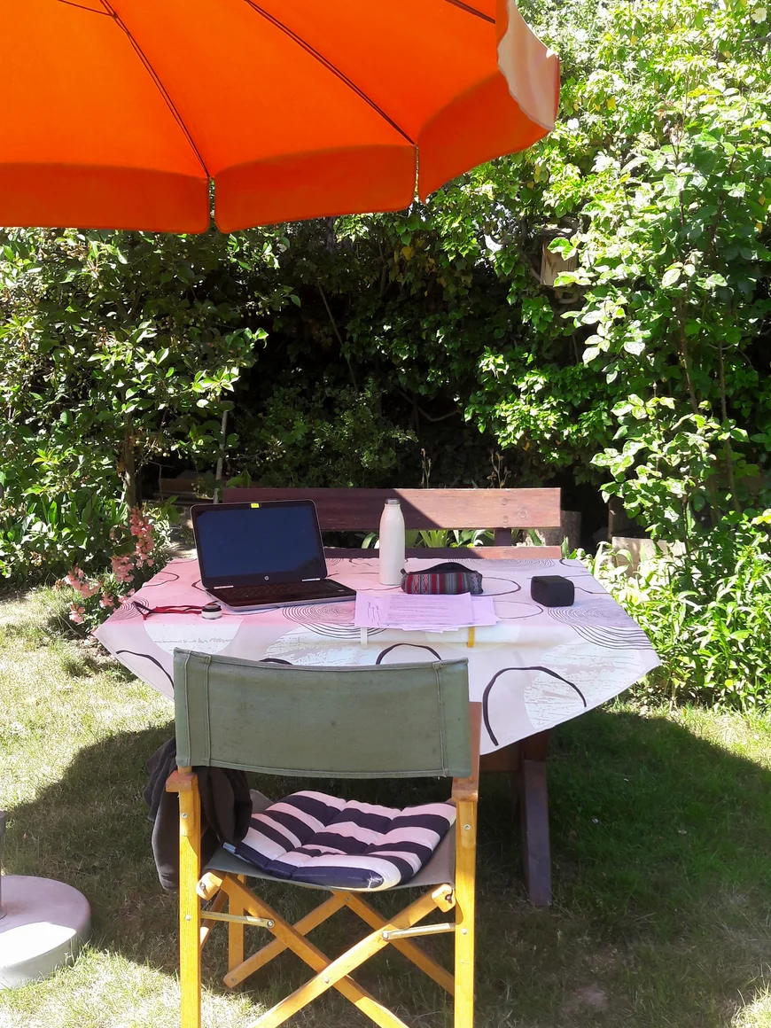In einem Garten unter einem organene Sonnenschirm steht ein GArtentisch mit Laptio und Schreibsachen