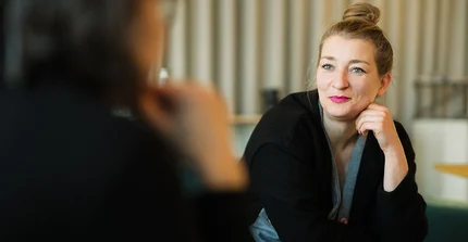 Foto von Dr. Anna Finzel mit sehr dunkler Kleidung und zusammengefassten Haaren, leicht vornübergebeugt im Gespräch