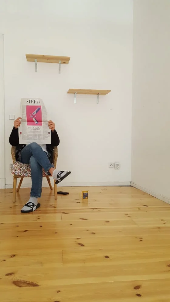 Ein leeres Zimmer. in der linken Ecke des bildes sitzt eine Person auf einem Korbstuhl und hölt eine Zeitung vor ihr GEsicht. Die Schlagzeile, die zu sehen ist, lautet "Streit". Auf dem Boden steht eine Kaffeetasse, an der Wand hitner der PErson hängen zwei leere Regalbretter