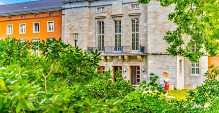 Durch Büsche gerahmter Blick auf das Portal eines großen Hauses, vor dem eine Rotkäppchenfigur steht