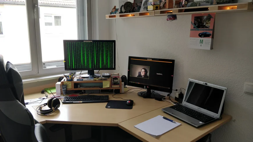 Eckschreibtisch. Drei Bildschirme, auf dem linken laufen grüne nullen und einsen auf schwarzem hintergrund von oben nach unten wie in dem FIlm Matrix. Auf dem Mittleren Videochat. Auf dem rechten nichts. Auf dem Tisch liegen Schreibutensilien, Kopfhöhrer, TAstatur, Mouse und ein Regal mit Krams